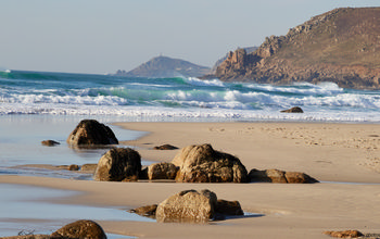 Cape Cornwall from Sennen beach This landscape photograph shows Cape Cornwall viewed from Sennen beach on the Cornish coast in the United Kingdom during a spring afternoon. The scene captures the natural beauty of Sennen Cove, with waves breaking onto the sandy beach and several large rocks scattered across the shoreline. The rugged cliffs of Cornwall rise in the background, adding depth to the image and highlighting the region’s distinctive coastline. Cape Cornwall, a well-known landmark, is visible in the distance, with its promontory and historic structure clearly identifiable. The combination of coast, nature, cliffs, and beach under soft spring sunlight illustrates the character of Cornwall’s western shores.